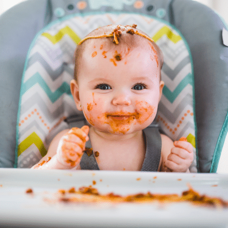 Bebé sonriente con la cara y las manos manchadas de comida mientras aprende a comer con el método Baby Led Weaning.