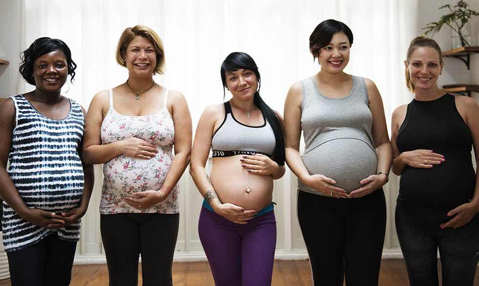 Grupo de cinco mujeres embarazadas de diferentes etnias sonriendo y posando juntas en una sala iluminada, con las manos sobre sus vientres. Dietista materno-infantil y asesora de lactancia en Sevilla